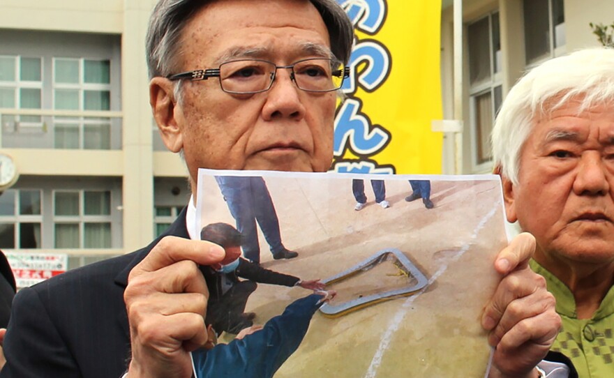 Okinawa Governor Takeshi Onaga, stands outside an elementary school as he shows a picture of the window fallen from U.S. military helicopter onto the sports ground of the school in Ginowan, Okinawa, on Wednesday.