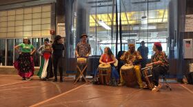 Four people sit with drums while third plays a maraca and two dancers prepare to perform.