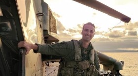 Maj. Tyler Braconi, in green flight suit, stands on the landing gear of a military helicopter smiling on the flight deck of a Navy ship.
