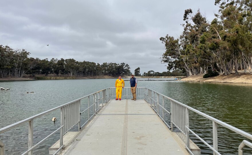 Two people stand on the edge of a pier at Chollas Lake, one wearing a bright yellow tracksuit.