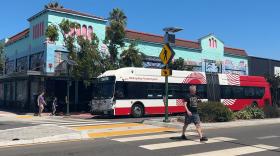 A pedestrian wearing a black shirt and grey shorts walks in the crosswalk on University Avenue in North Park. A red and white MTS is making a stop in the background in front of a bright blue building. The sky is clear and blue.