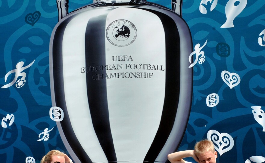 <strong>Larger Than Life:</strong> Tourists pose in front of a UEFA Euro 2012 Cup placard on Kiev's Independence Square in Ukraine. Europe is entering a packed sports schedule — but soccer still reigns supreme, says Frank Deford.