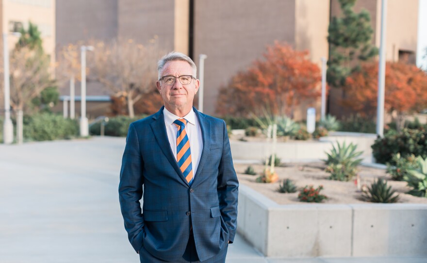 El Cajon Mayor Bill Wells stands with his hands in his pockets outside in a blue suit, white shirt and striped orange and blue tie.