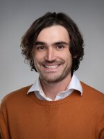 Man smiling with dark hair wearing an orange sweater.