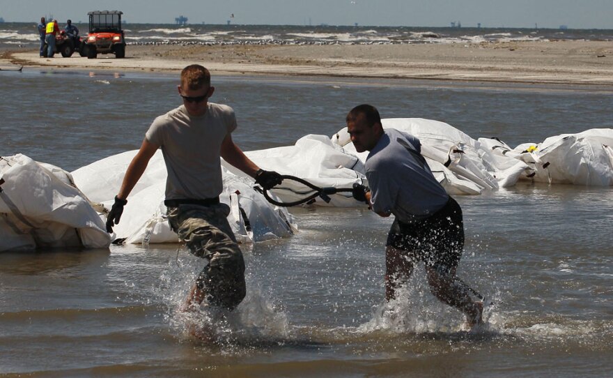Louisiana National Guard members retrieve a sling-load cable as helicopters drop sandbags into place in an estuary.