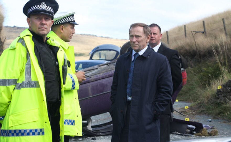 Chris Crowley (Danny Webb) stands next to the car crash with other police officers.