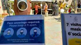 Patrons talk to an artist at the Sculptors Guild at Spanish Village Art Center in Balboa Park on Aug. 30, 2020, as a sign at the entrance tells visitors to keep 6 feet apart and wear their facial coverings to stem the spread of the coronavirus.