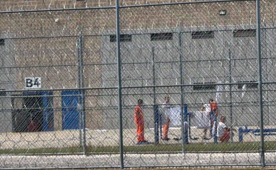 Detainees spend time outdoors behind barbed wire, as seen from a sandy public road that runs behind the facility.
