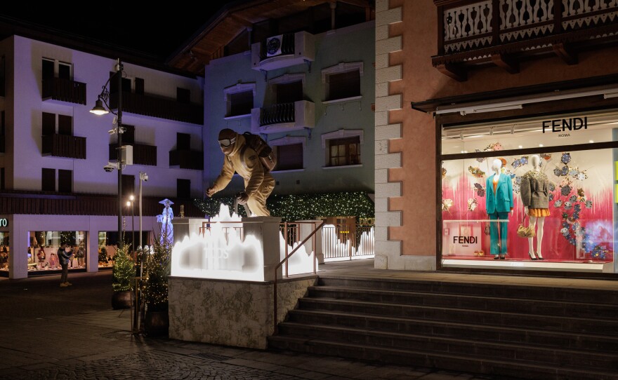 A window display for the luxury fashion brand Fendi illuminates a central street in Cortina d'Ampezzo, adjacent to a large outdoor sculpture of a skier. The town's main thoroughfare is a glamorous hub where premier designer brands like Dior, Gucci and Prada are represented.