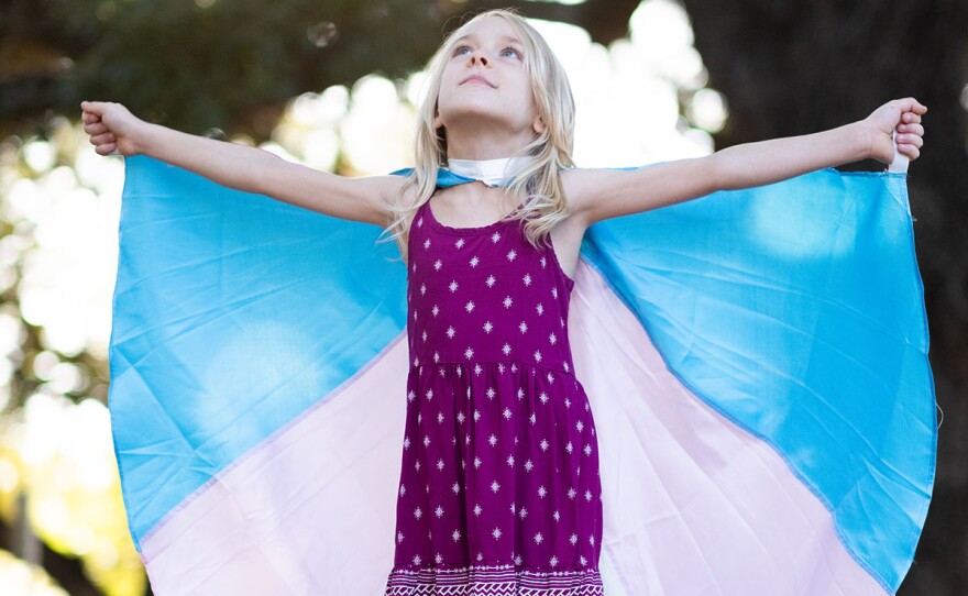 Kai looking up with her arms outstretched, wearing the trans flag like a cape