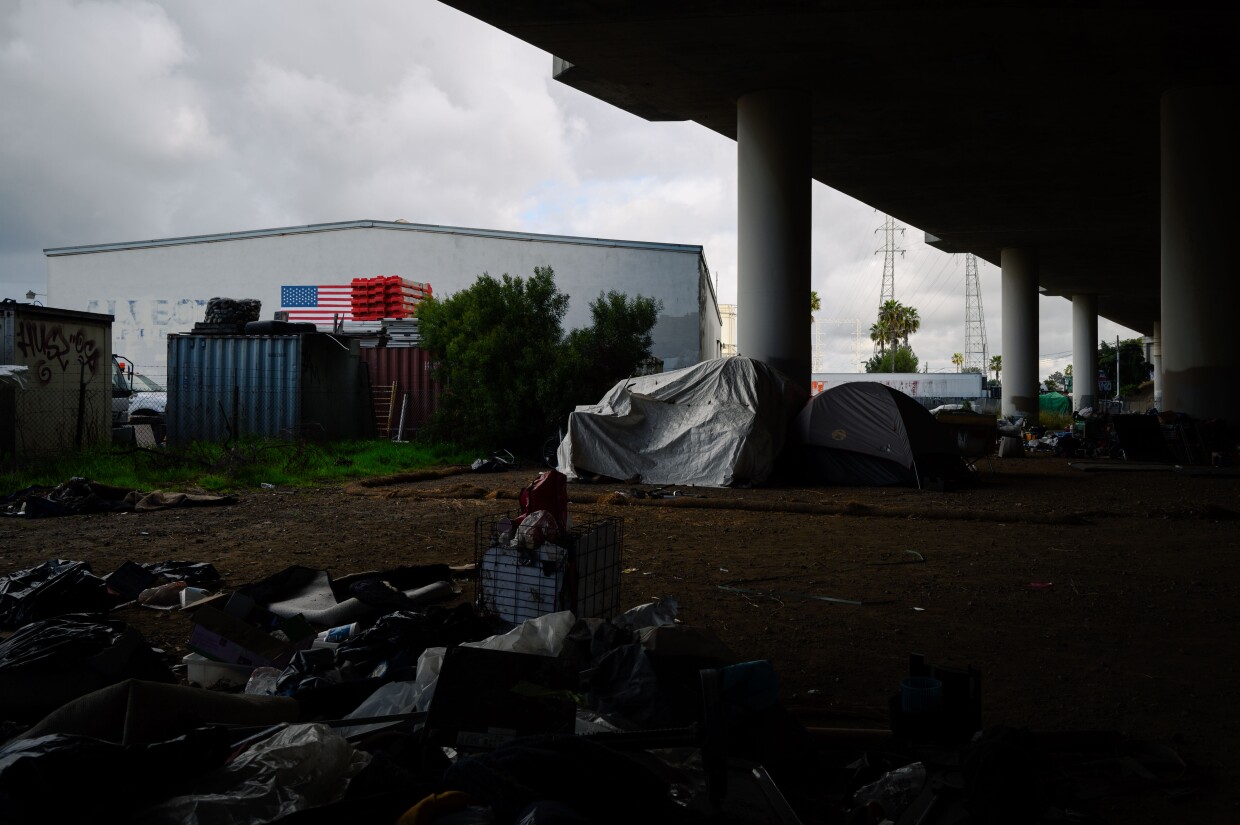 Tents and debris are seen beneath an American flag under an I-5 overpass in National City on March 7, 2024. National City is the second in San Diego County to move away from a police-led response to homelessness and toward trained caseworkers.