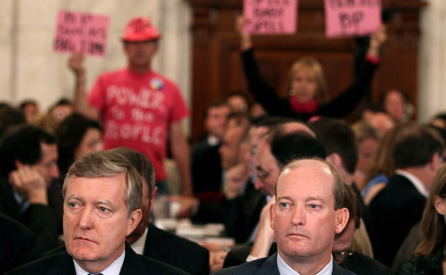 BP America's Lamar McKay and Executive Vice President David Nagel wait to testify as people hold signs of protest behind them.