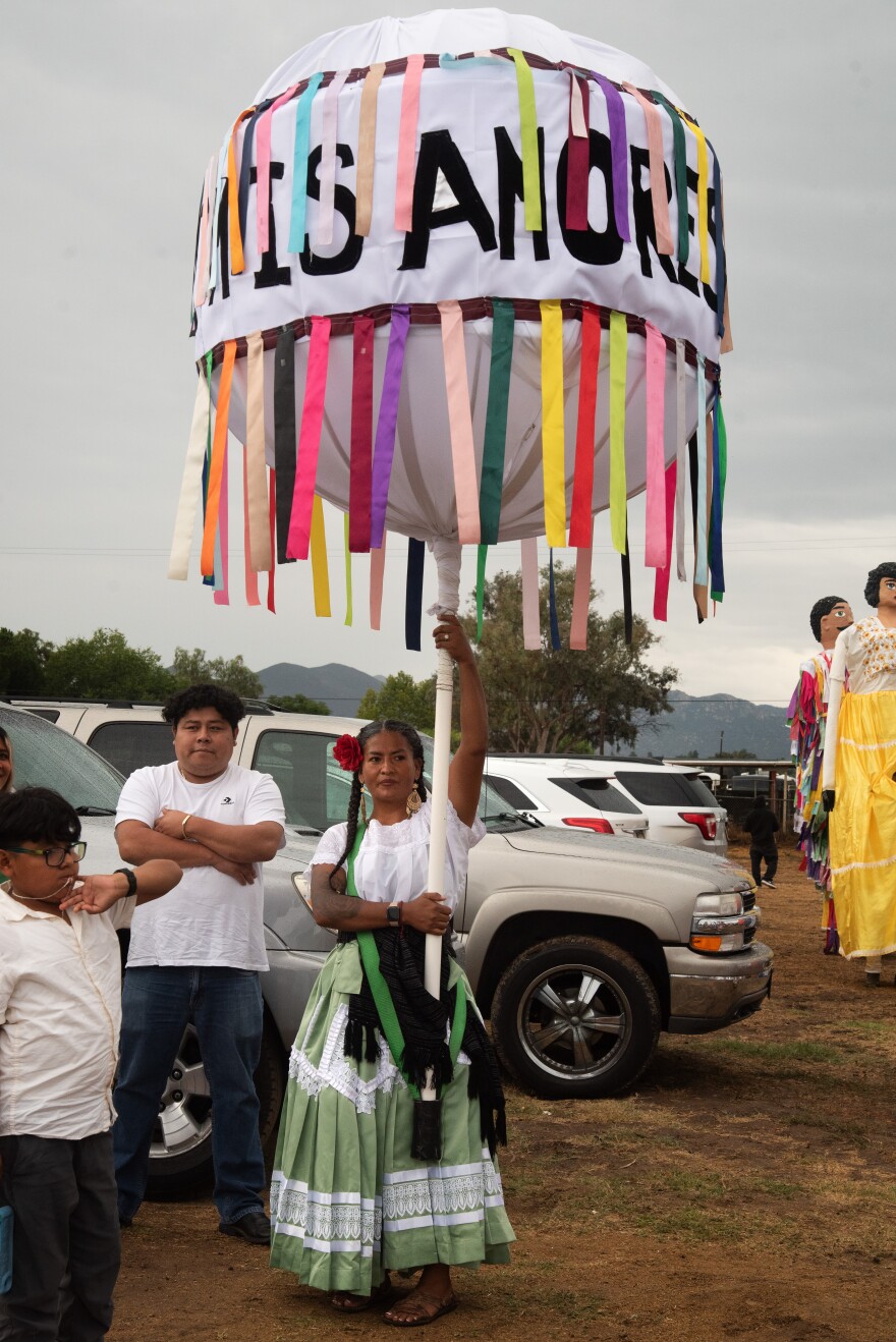 Rosie Molano holds a giant large white balloon called a marluta, adorned with colorful ribbons and the words ‘Candela Valles Centrales’ and ‘Oaxaca de Mis Amores’ before her dance group performs at a event in Valley Center on September 21, 2025. Molano leads the Oaxacan dance group Candela Valles Centrales with her husband, who also carries a marluta.