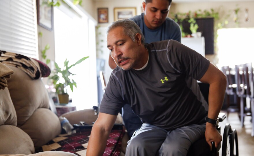 Joel Ramirez climbs back into his wheelchair with the help of Francisco Guardado, a home health aide, at his home in Rialto, Calif.