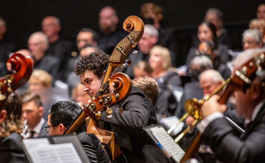 Performers in the La Jolla Symphony and Chorus are shown in an undated photo. A man playing double bass is standing in the center of the photo, leaning forward and concentrating on the music. The other musicians and the choir are not in focus.
