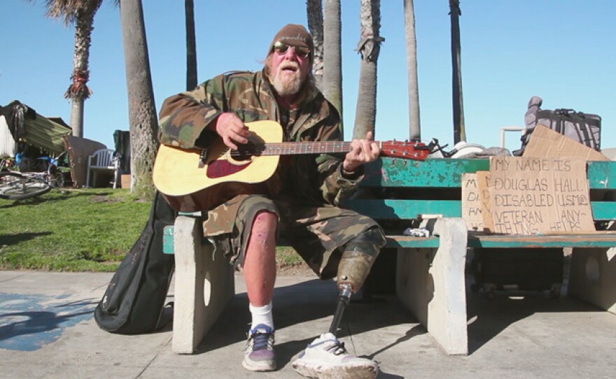 A disabled man sits on a bench playing guitar with a sign that reads "My name is Douglas Hall, disabled U.S.M. veteran - anything helps" in Venice, Calif. Homelessness and poverty are major issues explored in ONE DAY IN THE AMERICAN CITY.