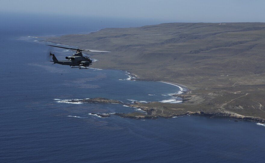 A Marine Corps Ah-1Z SuperCobra attack helicopter flies from the left toward the western rocky shore of San Clemente Island.