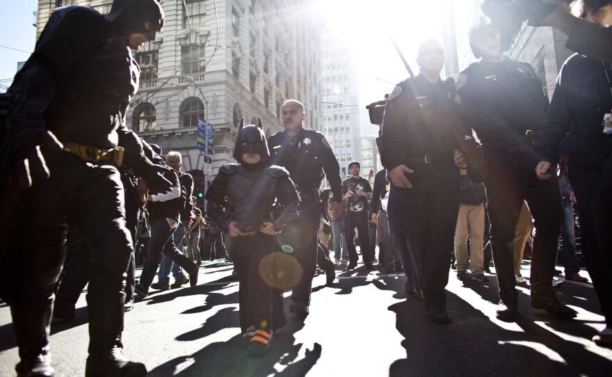 Police escort Batkid and Batman after they arrest The Riddler.