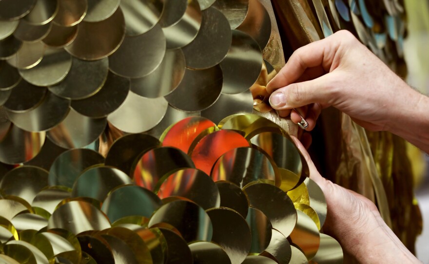 Costume designer Matthew Flower, who goes by Machine Dazzle, adjusts a gold cape worn by Belgian tenor <a href="https://www.clbmanagement.co.uk/melnik-maxime">Maxime Melnik</a> in his role as Apollo, one of two gods who tries to seduce titular character Io.