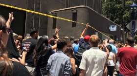 People walk under police tape after a San Diego police officer accidentally shot himself in the leg while pursuing a hit-and-run suspect who pointed a weapon at police and was eventually arrested on the roof of a parking structure near the finish line of an annual marathon, authorities said, Sunday, June 3, 2018, in San Diego.