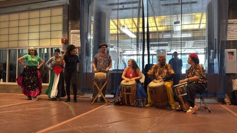 Four people sit with drums while third plays a maraca and two dancers prepare to perform.