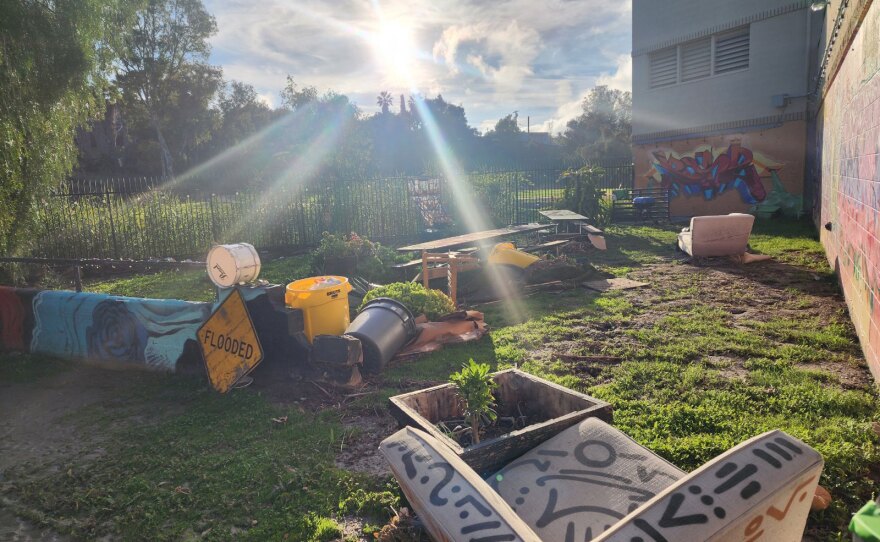 Garden equipment, plants and furnishings are strewn about an outdoor space. Graffiti murals adorn the walls. The area looks muddy. A yellow diamond "FLOODED" sign is propped against some debris.