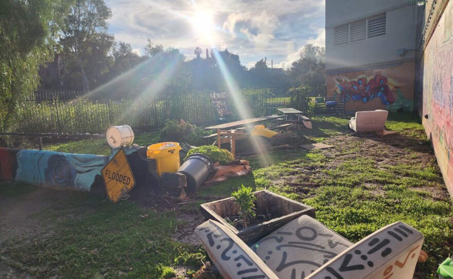 Garden equipment, plants and furnishings are strewn about an outdoor space. Graffiti murals adorn the walls. The area looks muddy. A yellow diamond "FLOODED" sign is propped against some debris. 