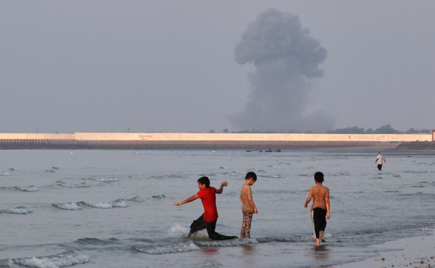 Three boys play in the shallow waters of the Strait of Hormuz, as a plume of smoke rises from an explosion in the background, off Bandar Abbas, Iran, Monday.