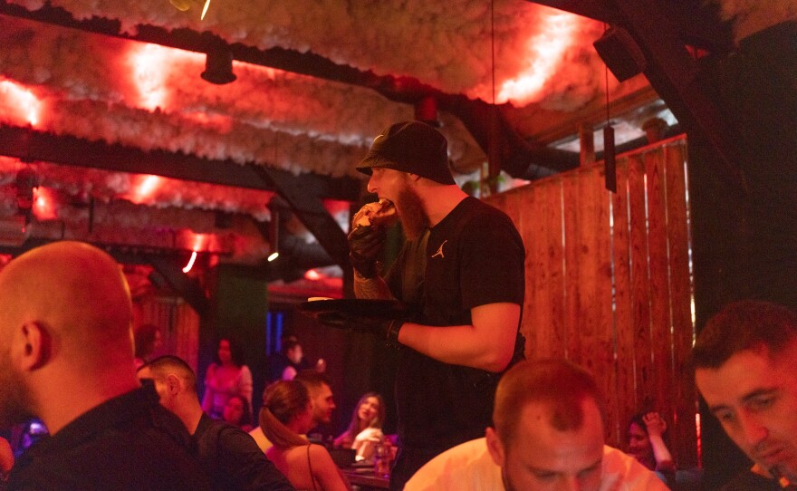 A man eats a burger as he walks to his table, wearing gloves to keep his hands clean.
