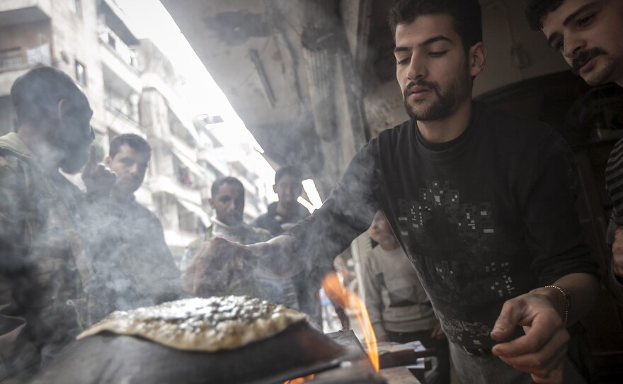 A man makes bread as residents, background, stand in line in front of a bakery during heavy fighting between Free Syrian Army fighters and government forces in Aleppo, Syria, on Dec. 4, 2012.