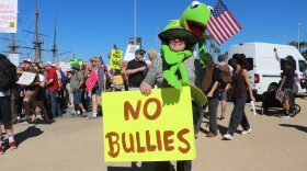 Mary Decker is wrapped with a Kermit the Frog plush toy and holds a sign that reads "No Bullies" during the "No Kings" protest rally at Waterfront Park on Oct. 18, 2025.