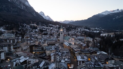 The illuminated bell tower of the Basilica Minore dei Santi Filippo e Giacomo stands at the heart of Cortina d'Ampezzo, Italy, as evening settles over the valley. Once a small village of farmers and shepherds, this storied town has evolved into the "Pearl of the Dolomites," a renowned luxury destination. Surrounded by the limestone peaks of the UNESCO World Heritage Dolomites, the town's historic center remains a "living room" for celebrities and high society.