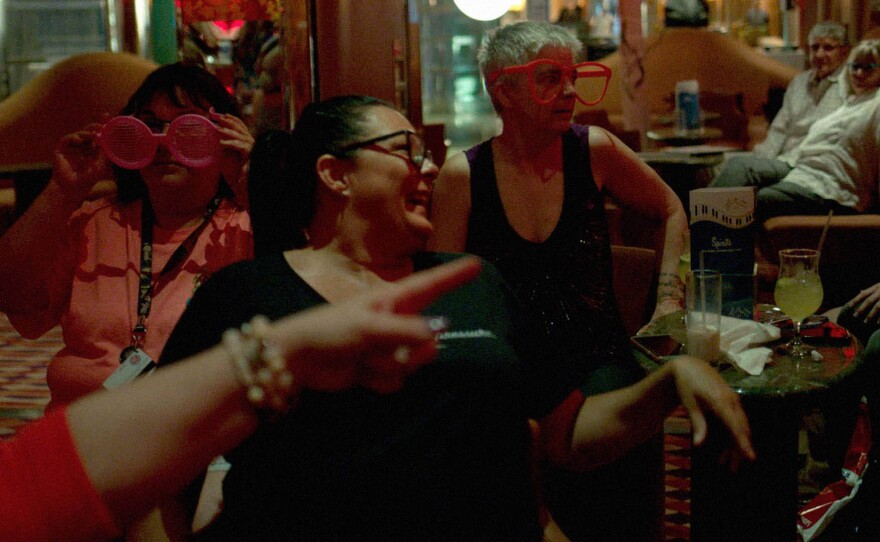 The film’s protagonist, Desiree Wood, a Mexican-American woman wearing a black t-shirt and glasses with hair pulled back in a ponytail, seated with a group of friends in a dark bar with red-hued lighting looking to her left and laughing, flanked by a brunette white woman in a bright pink t-shirt looking straight ahead and holding a pair of pink oversized plastic toy glasses in front of her eyes, and a white woman with cropped white hair and a loose purple sleeveless shirt looking to the left with a pair of oversized red-framed glasses, and in the foreground, a hand with its owner off-screen, pointing towards the left in the direction that Desiree is laughing.