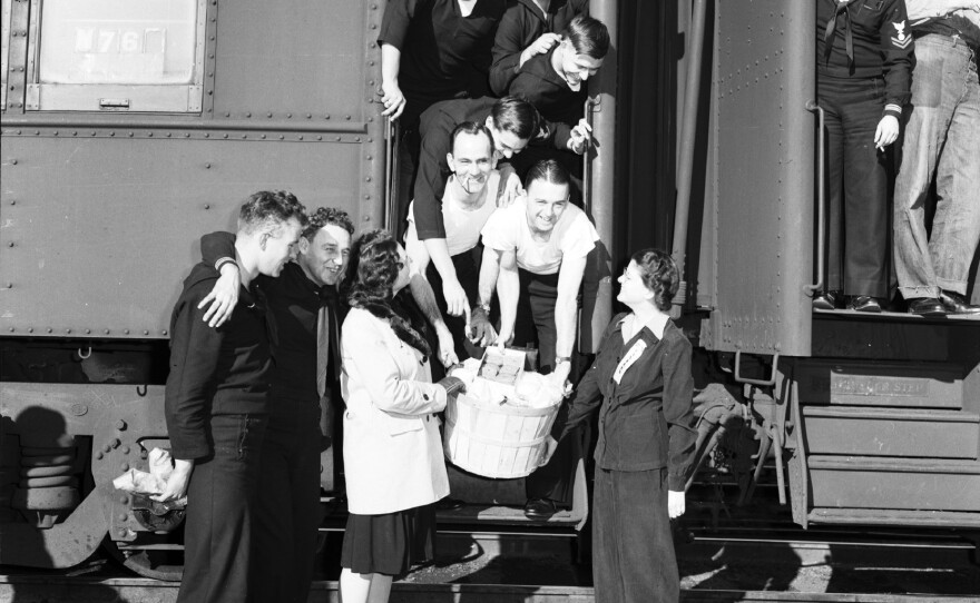 Soldiers pile into the doorway of a troop train to receive a basket from two canteen volunteers.