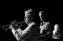 Gilbert Castellanos is shown in profile, playing trumpet, in a black and white photo with a black bacground. Out of focus in the background, other musicians look on.
