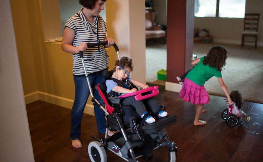 Kathleen helps Gideon into his chair while his sister Genevieve plays with her dolls, one of whom has a wheelchair.