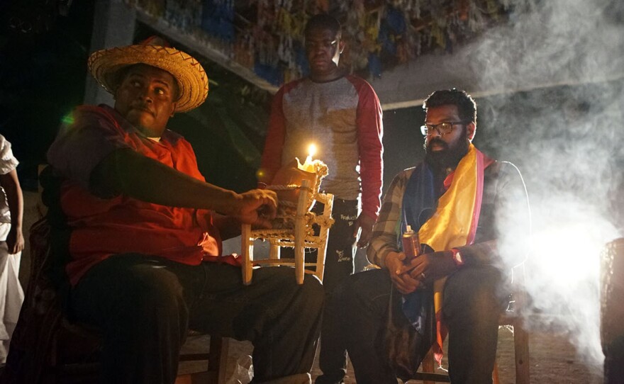 (R) Voodoo Priest (L) and Jeremy Dupin (standing) with Romesh Ranganathan in Haiti