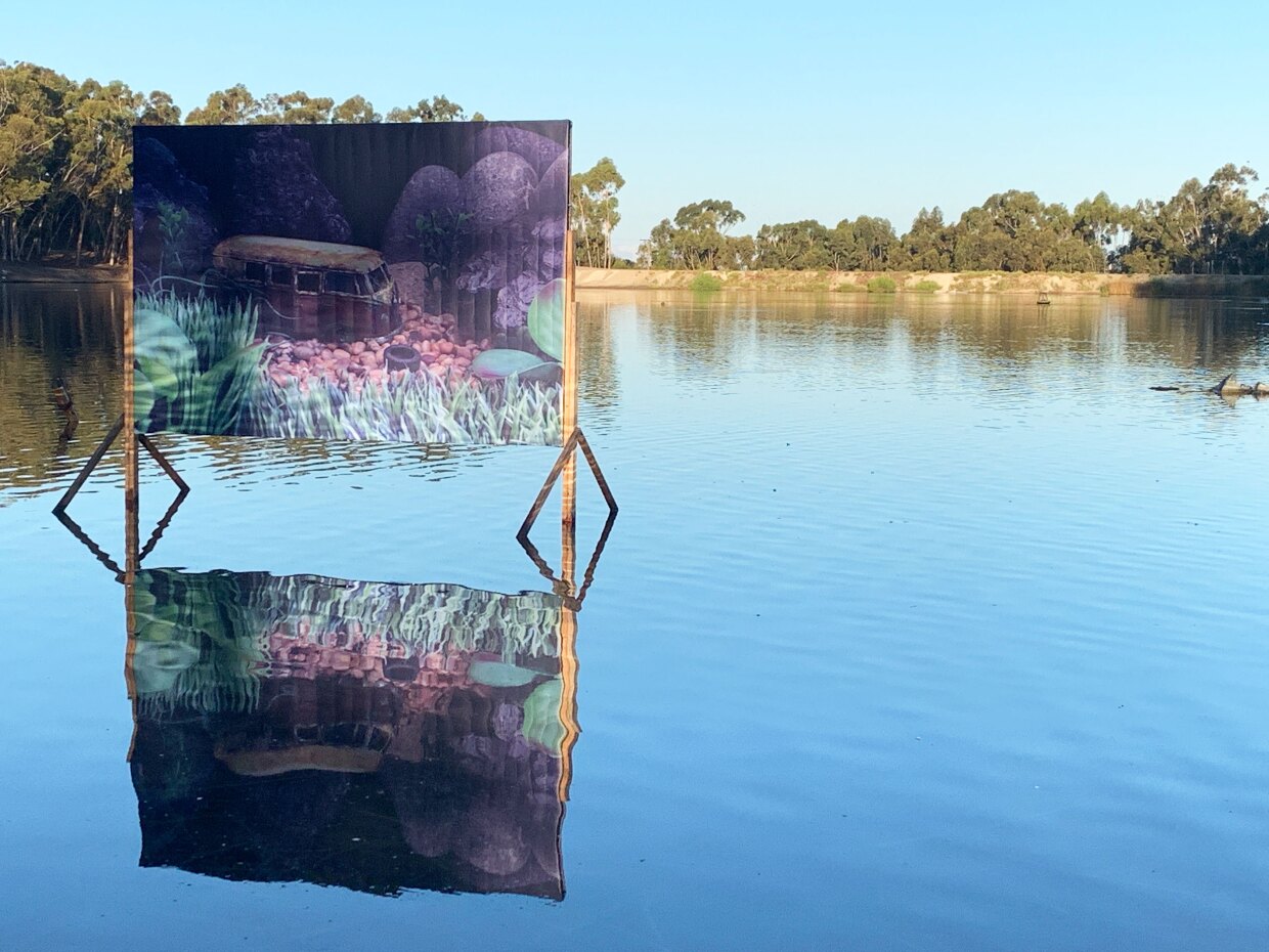 A billboard sits in a small lake, featuring a colorful, underwater scene with a painted image of a shipwrecked VW bus at the bottom of the lake. The billboard's reflection ripples on the lake's surface.