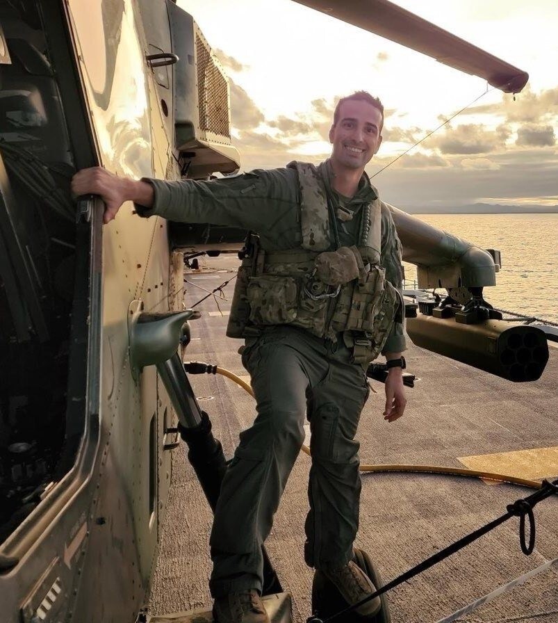 Maj. Tyler Braconi, in green flight suit, stands on the landing gear of a military helicopter smiling on the flight deck of a Navy ship.