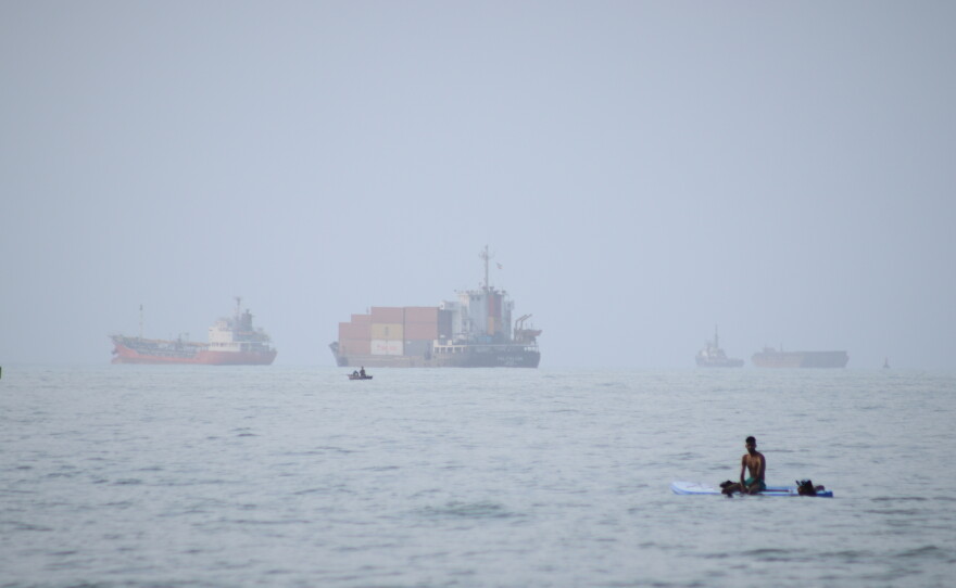 A man sits on a surfboard as a container ship and other commercial vessels appear anchored in the Strait of Hormuz off Bandar Abbas, Iran, Monday, July 27, 2026.