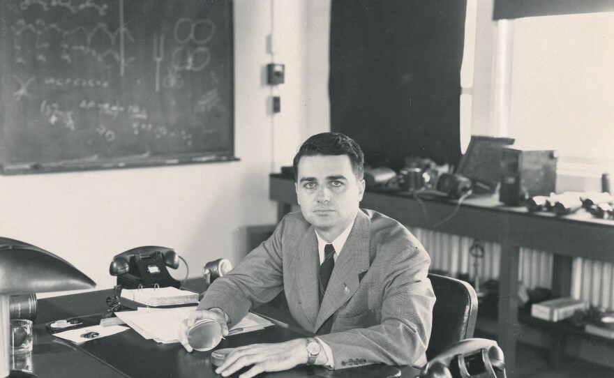 Edwin Land at desk holding circular object looking at camera.