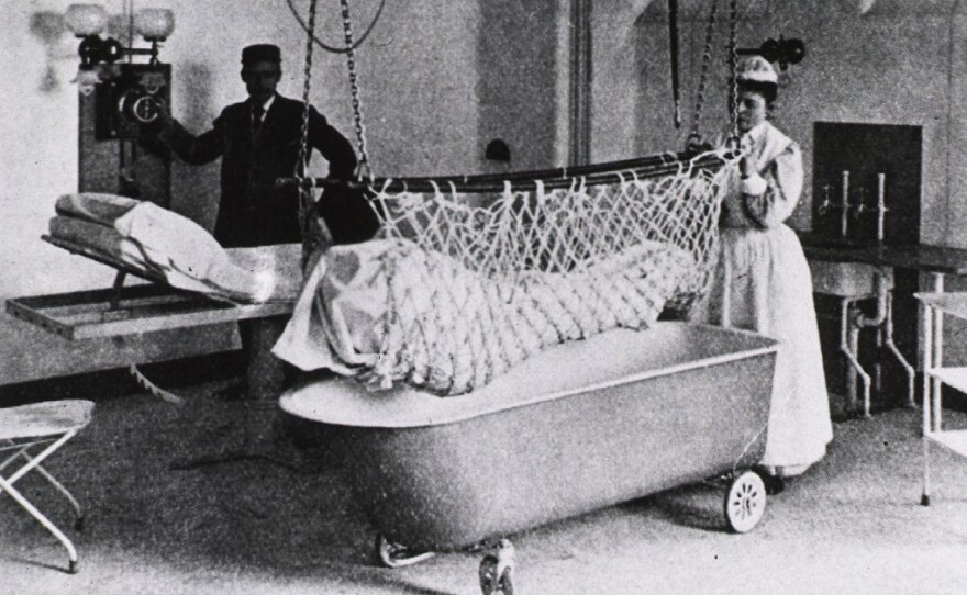 A nurse and an orderly move a patient from his bed to a bathtub with the aid of an "electric lift" in 1898.