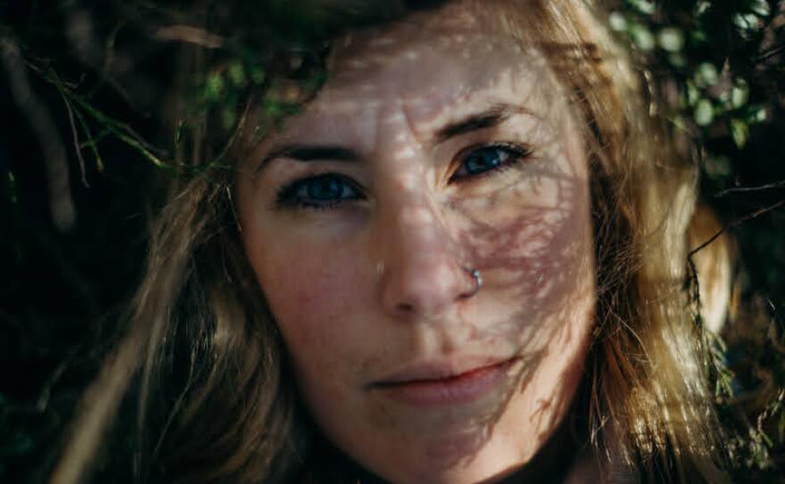 Maria's face is shown in close up and is framed by green leaves. She has blond hair, a nose ring in her right nostril and blue eyes. She stares straight ahead with a plaintive gaze. Shadows of branches are cast on right side of her face. Credit: 