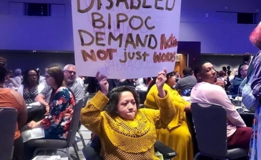 Sandy Ho holdings up a sign that reads "Disabled BIPOC demand actions not just words" in the middle of a conference room surrounded by many people sitting at tables.