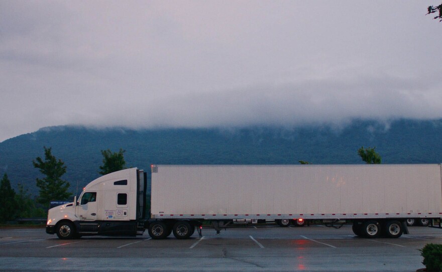 Long-shot of a white 18 wheeler with the film’s protagonist, Desiree Wood, in silhouette at the wheel, marker lights on, parked in an empty Walmart parking lot at dusk, wet concrete, green trees and blue mountains with green flora in the background, and fog hanging above the truck and covering the peak of the mountains along with a gray sky.