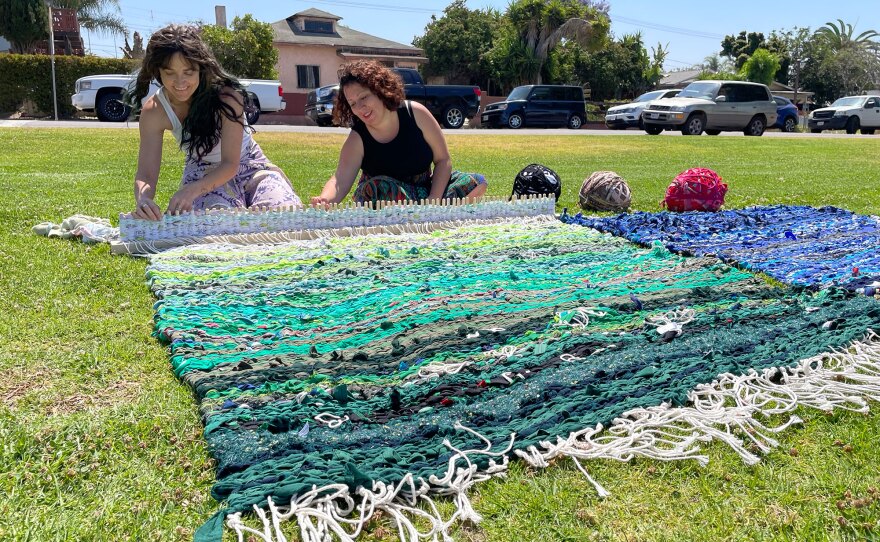 artists weave large, colorful blankets out of strips of fabric in a grassy park
