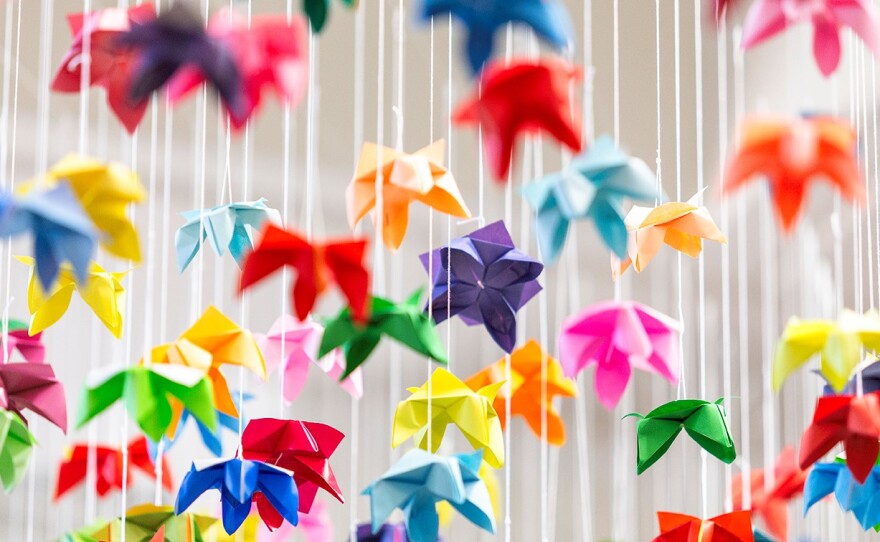 Dozens of differently colored origami shapes, which are shaped a bit like falling leaves, hang suspended from the ceiling, close together on white string. Colors include yellow, orange, bright green, bright purple, blue, light blue, and pink.