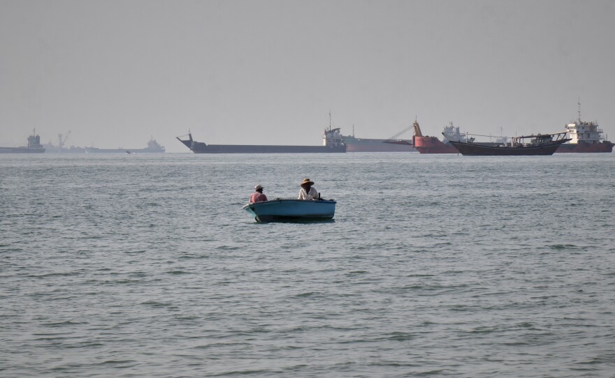 Two men sit in a small boat on the water as a mix of bulk carriers, cargo ships, and service vessels line the horizon in the Strait of Hormuz off Bandar Abbas, Iran, Monday, April 27, 2026.