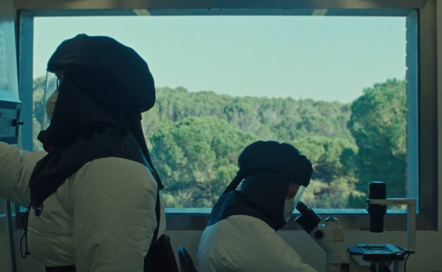 Two individuals wearing full hazmat suits work back to back in a laboratory. One stands and the other sits at a desk, looking through a microscope. Behind them, a large window stretching the width of the image shows a brilliantly green forest and blue sky outside the laboratory.