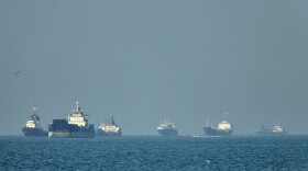 Cargo ships, including bulk carriers and general cargo vessels, sit at anchor offshore as a small motorboat passes in the foreground, in the Strait of Hormuz off Bandar Abbas, Iran, Monday, May 4 , 2026.